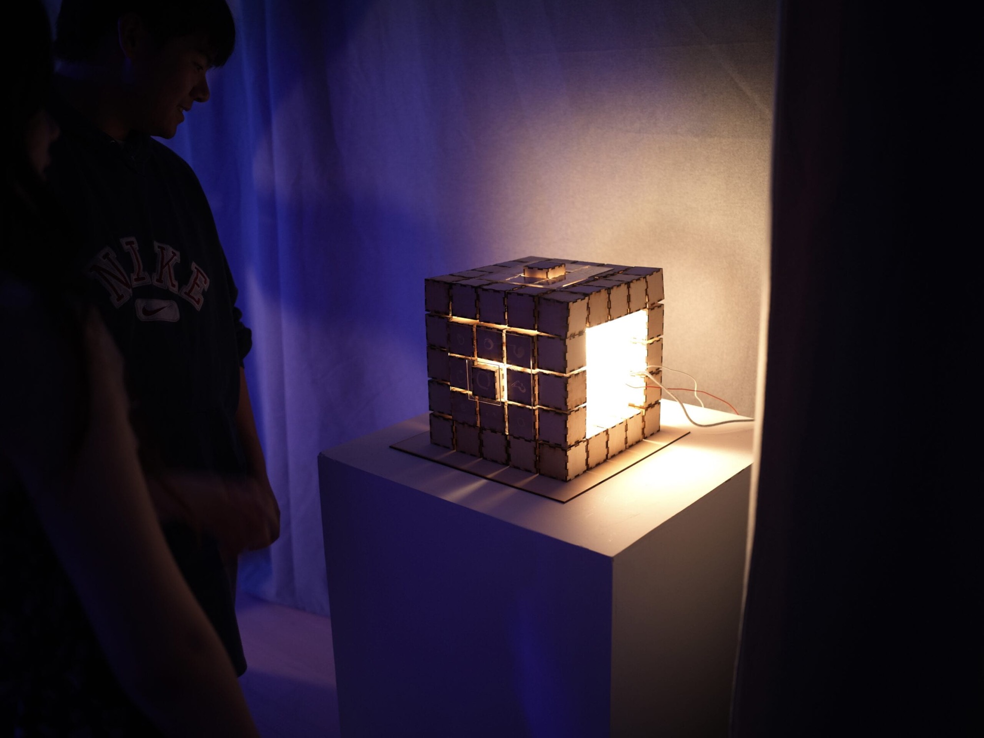 Evolving Spectrum on a white plinth at the IMA show, glowing warmly against blue-lit drapery as a visitor looks on